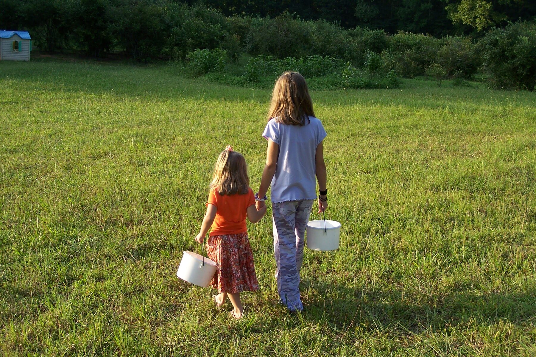 Two children walking hand in hand across the grass carrying white picking buckets